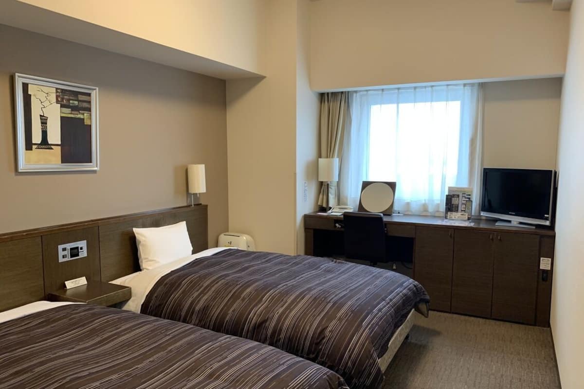A hotel room with two beds, a desk with a computer, a TV, and a window with curtains.