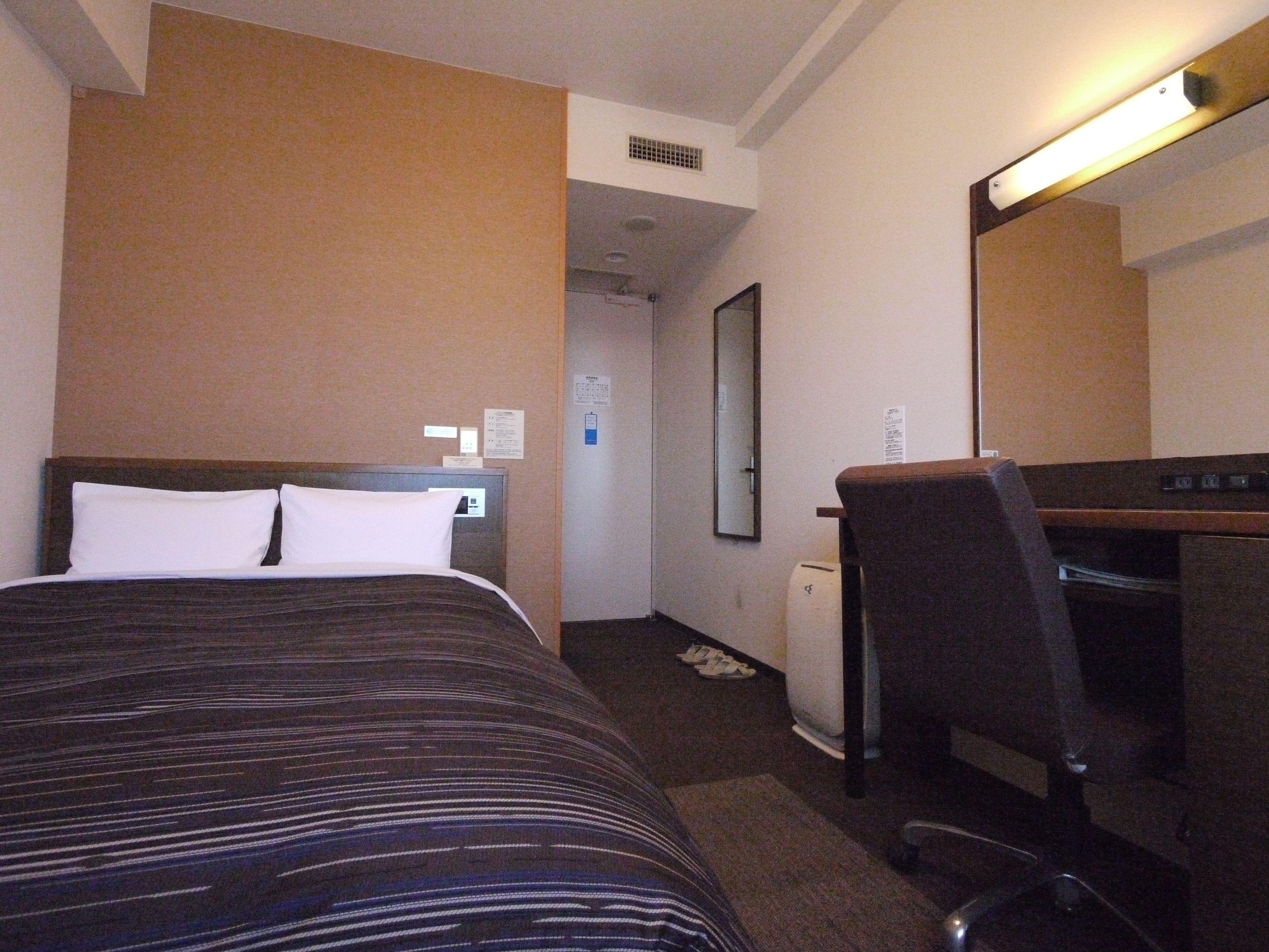 Una habitación de hotel con cama, escritorio, silla, espejo y una kitchenette pequeña.