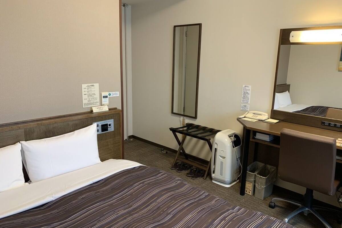 A hotel room with a bed, a desk with a chair, a mirror, a shoe rack, and a trash bin.