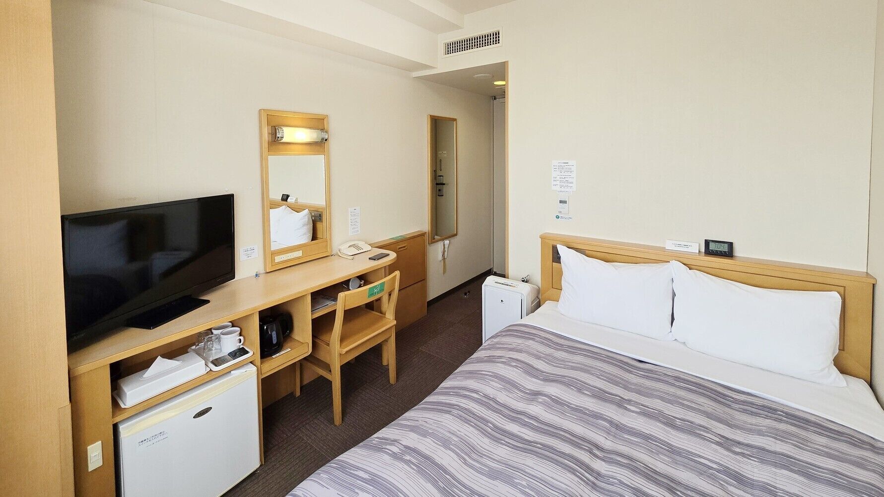 A hotel room with a bed, a desk with a television, a small refrigerator, and a mirror.
