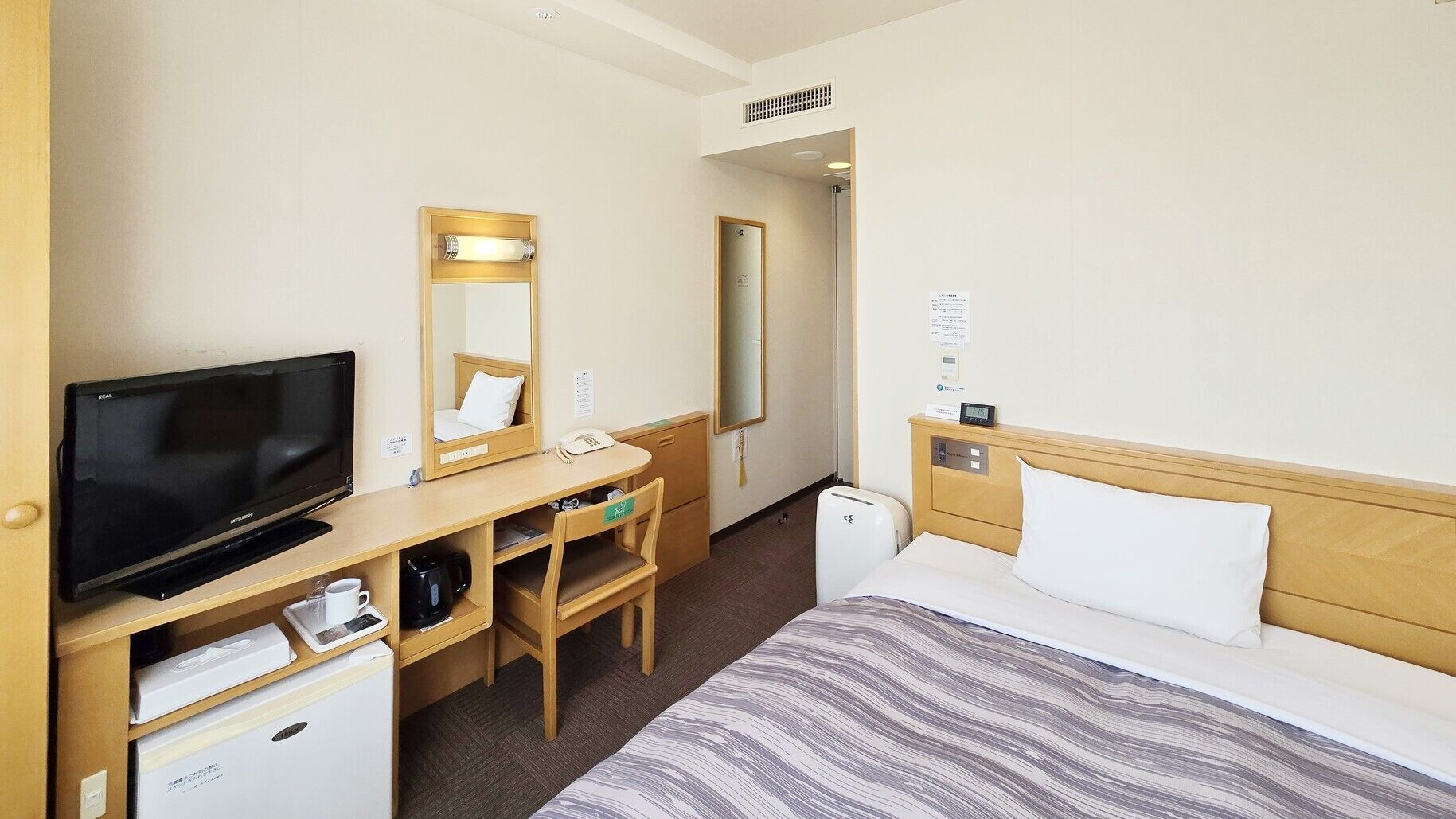 A hotel room with a bed, a desk with a computer, a television, a mirror, and a small refrigerator.