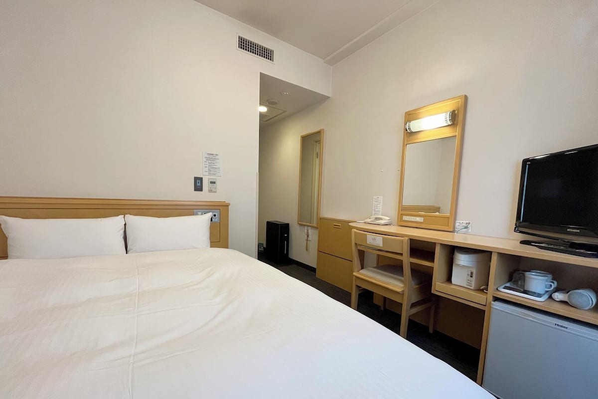 A hotel room with a bed, a desk with a mirror, a television, and a small refrigerator.