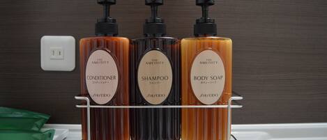 Un baño con un estante que contiene tres botellas de productos de tocador de la marca The Amenity: acondicionador, champú y jabón corporal.