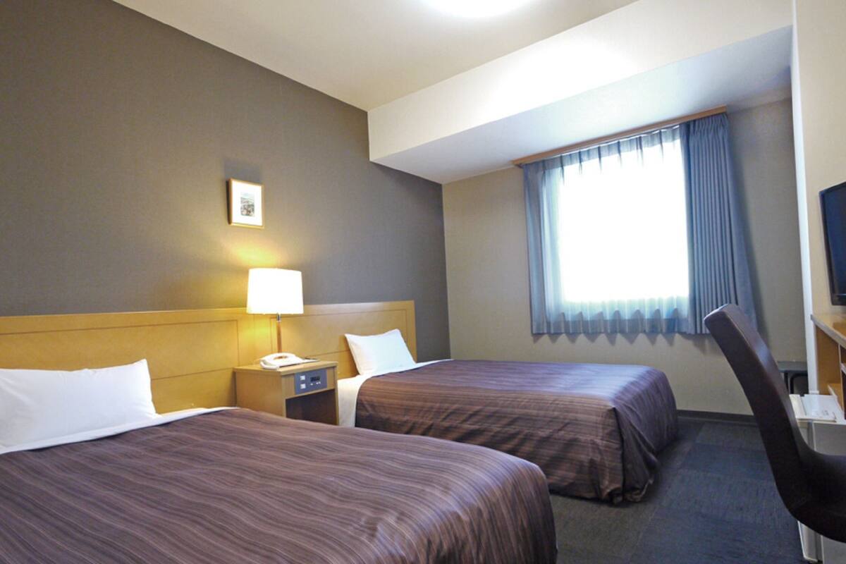 A hotel room with two beds, a desk, a chair, a TV, a lamp, and a window with curtains.