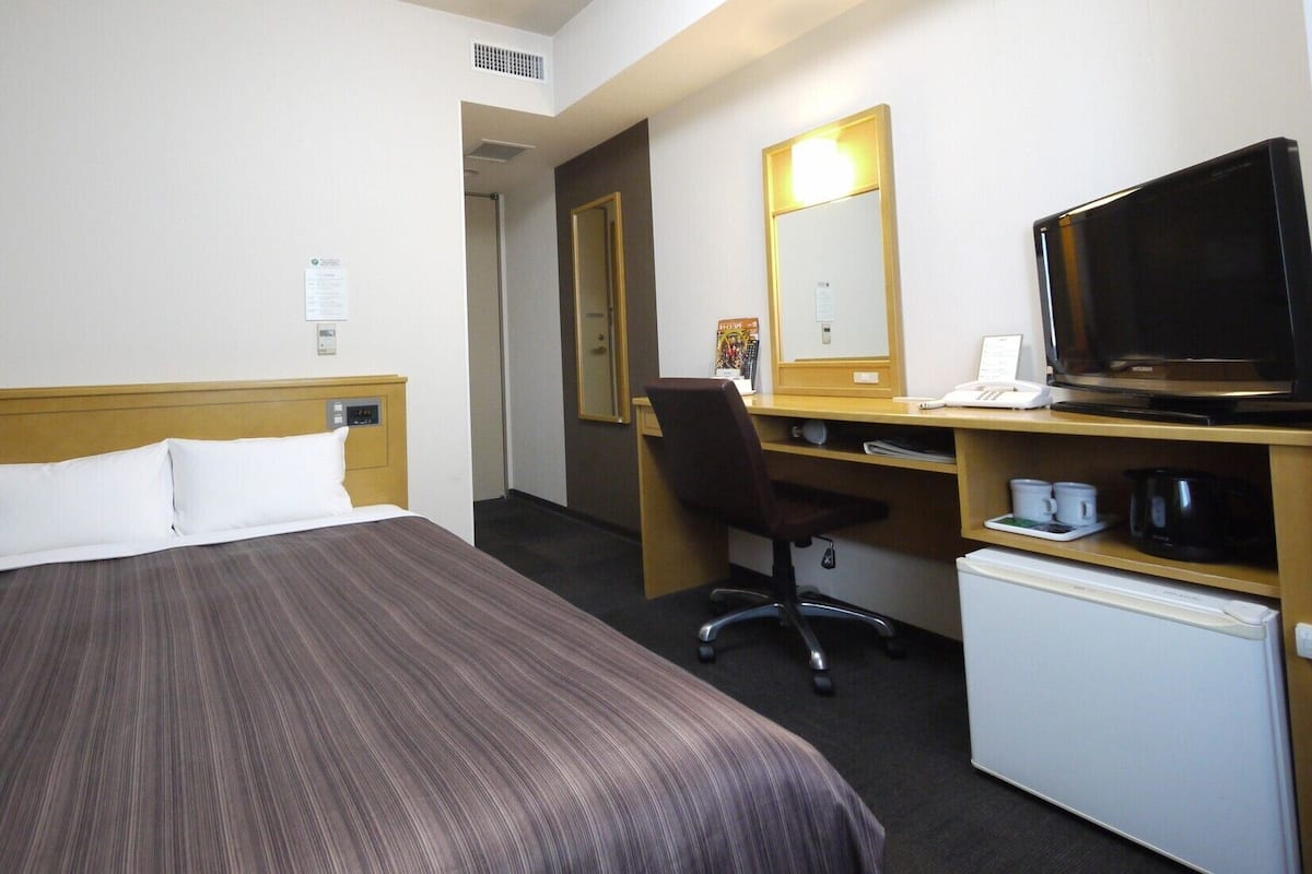 A hotel room with a bed, a desk with a computer, a television, and a mini-fridge.