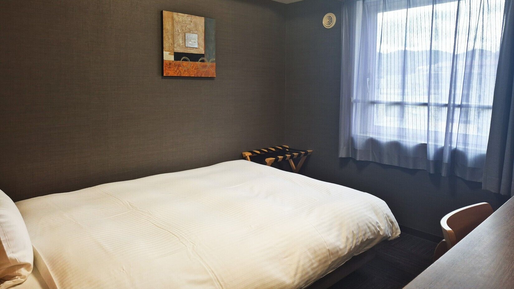 A hotel room with a bed, a chair, a small table, a painting on the wall, and a window with curtains.