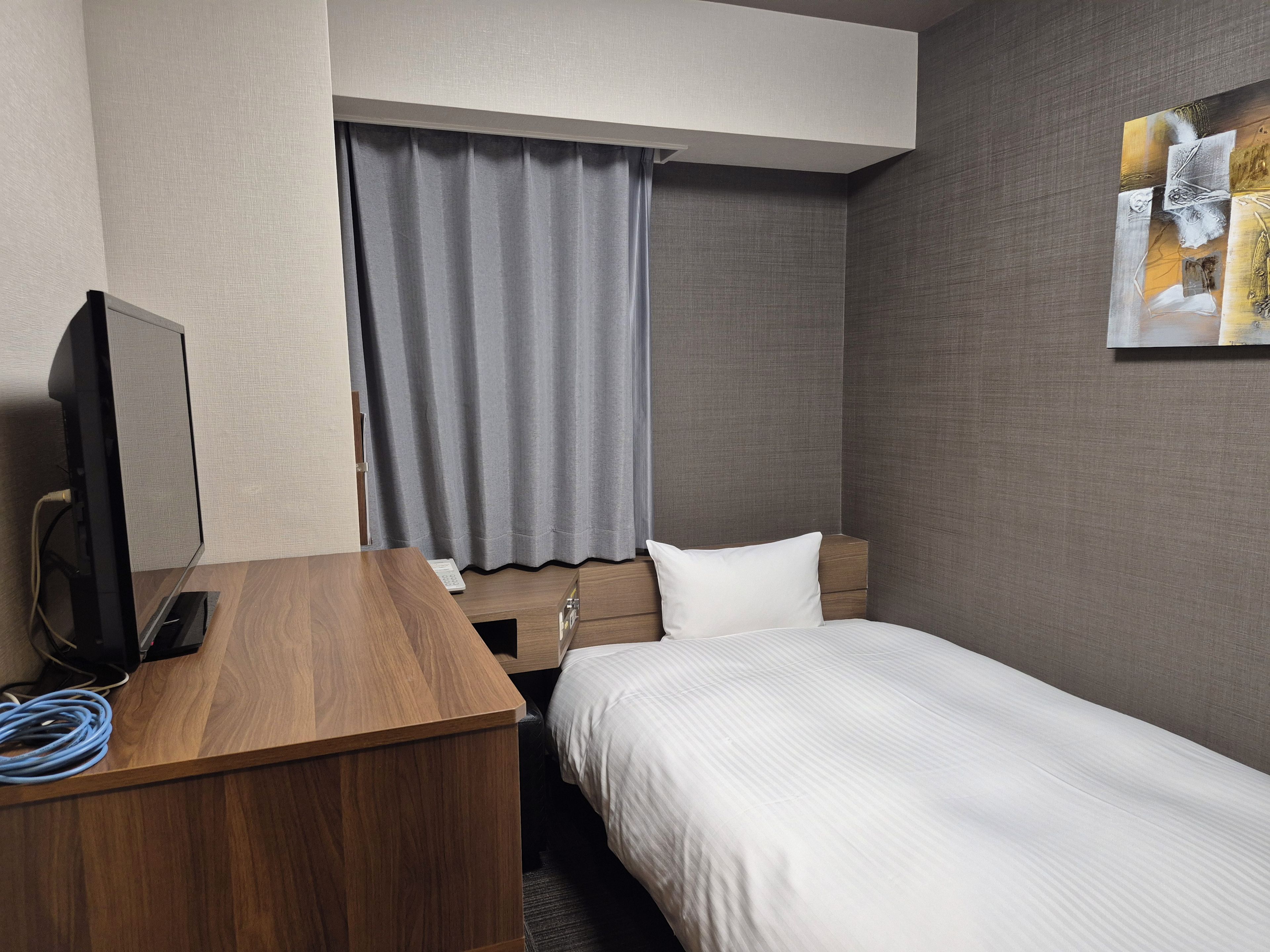 A hotel room with a wooden desk, a television, a bed with white bedding, a grey curtain, and a painting on the wall.