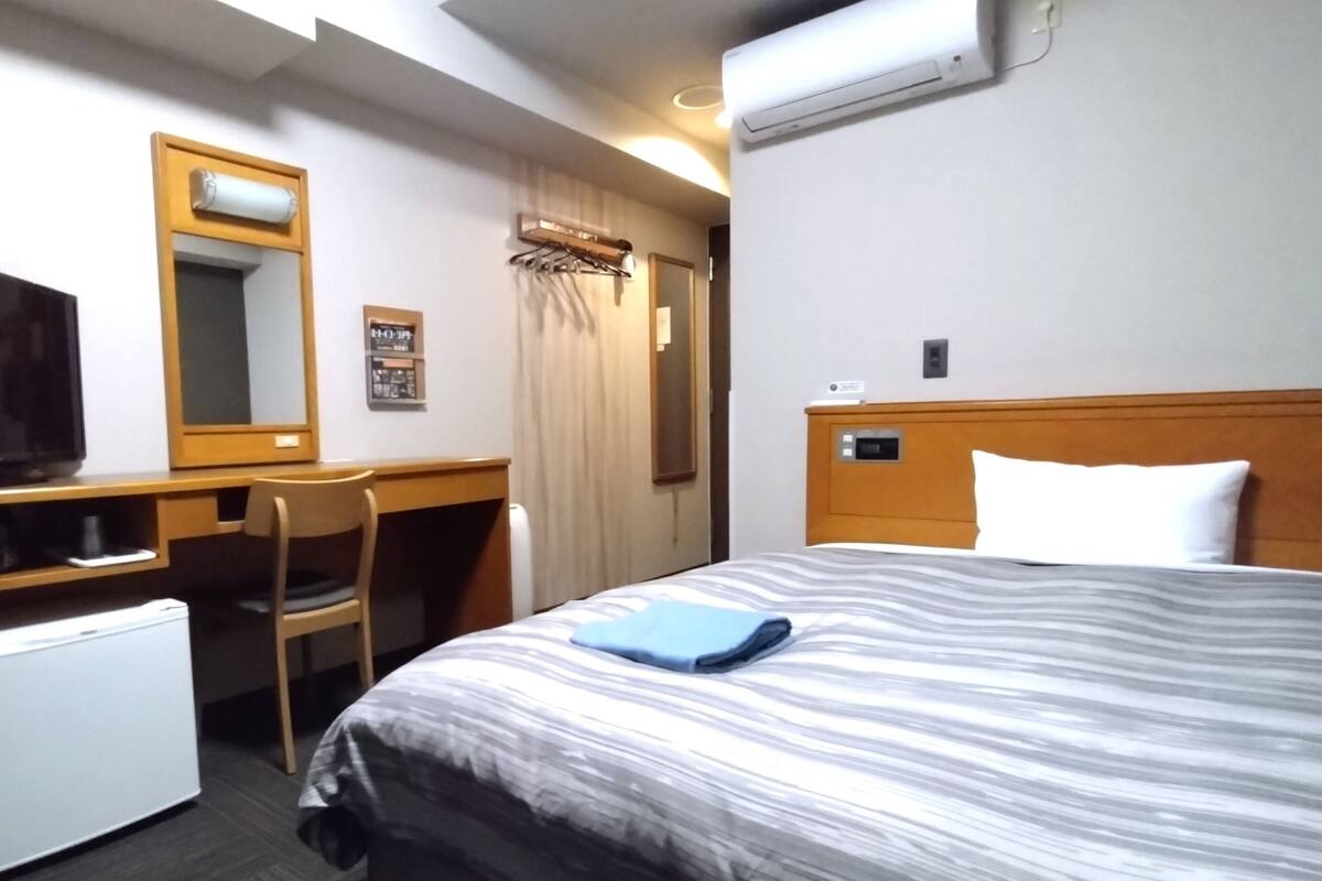 A hotel room with a bed, a desk, a chair, a television, and an air conditioning unit.