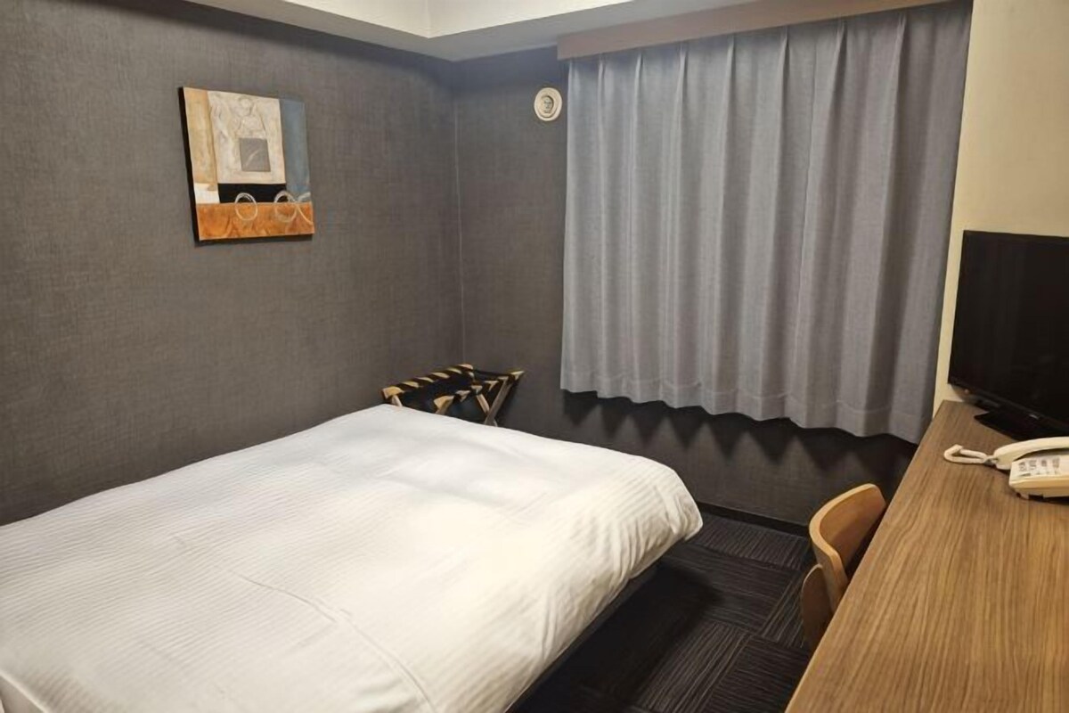 A hotel room with a bed, a desk with a telephone, a chair, a window with curtains, and a wall-mounted artwork.