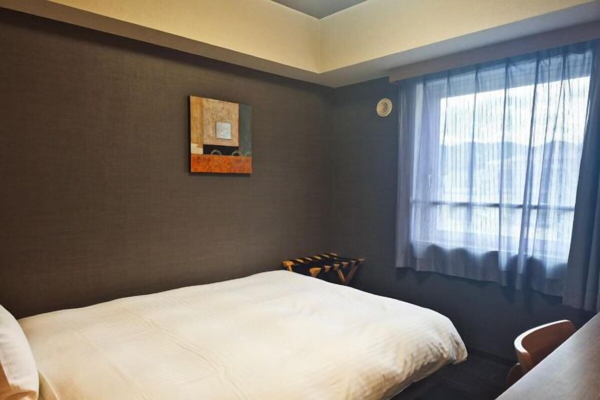 A hotel room with two beds, a window with curtains, a wall clock, and a small table with a chair.