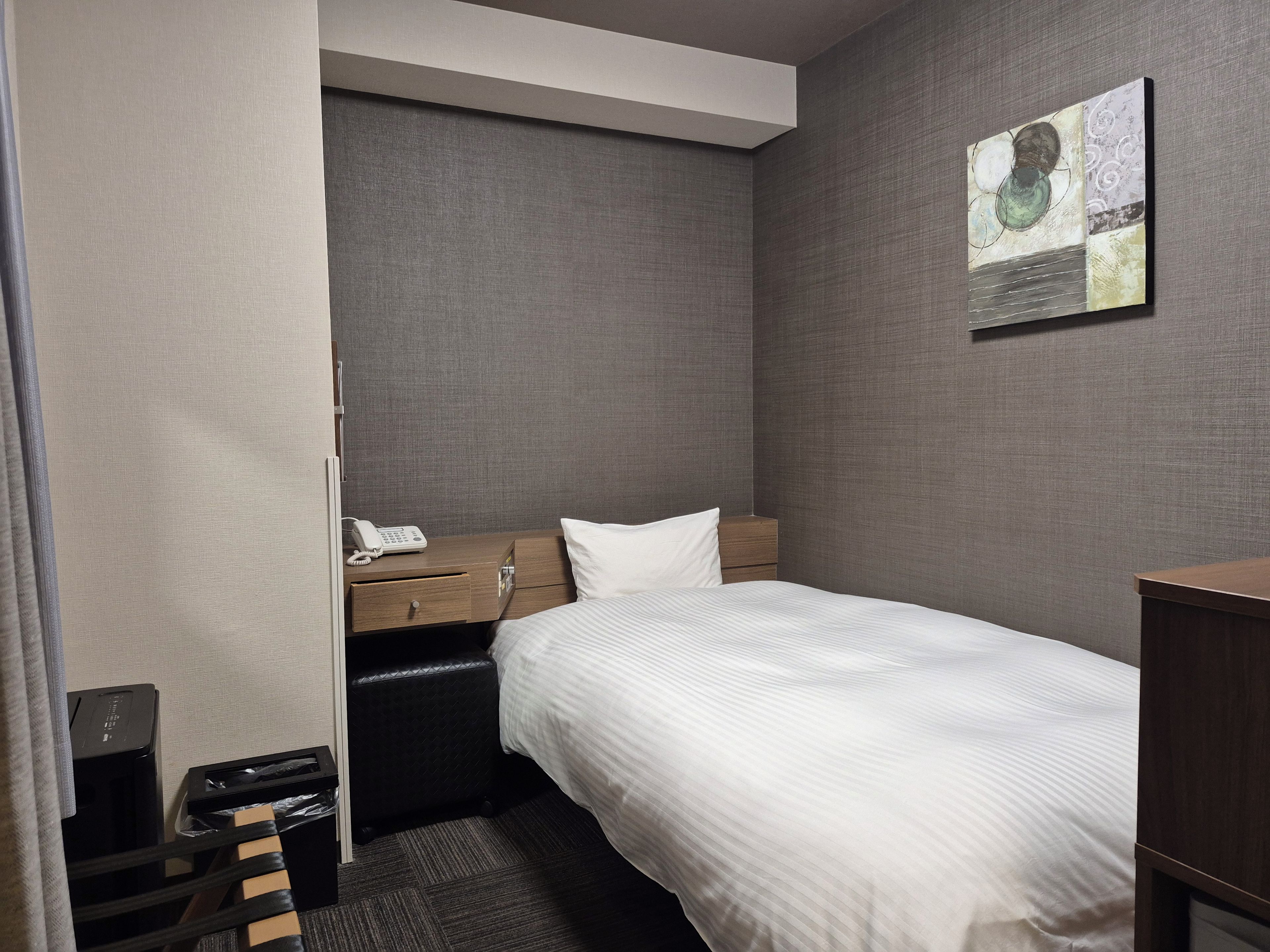 A hotel room with a single bed, a wooden nightstand, a telephone, a black leather chair, a small table, and a wall-mounted artwork.