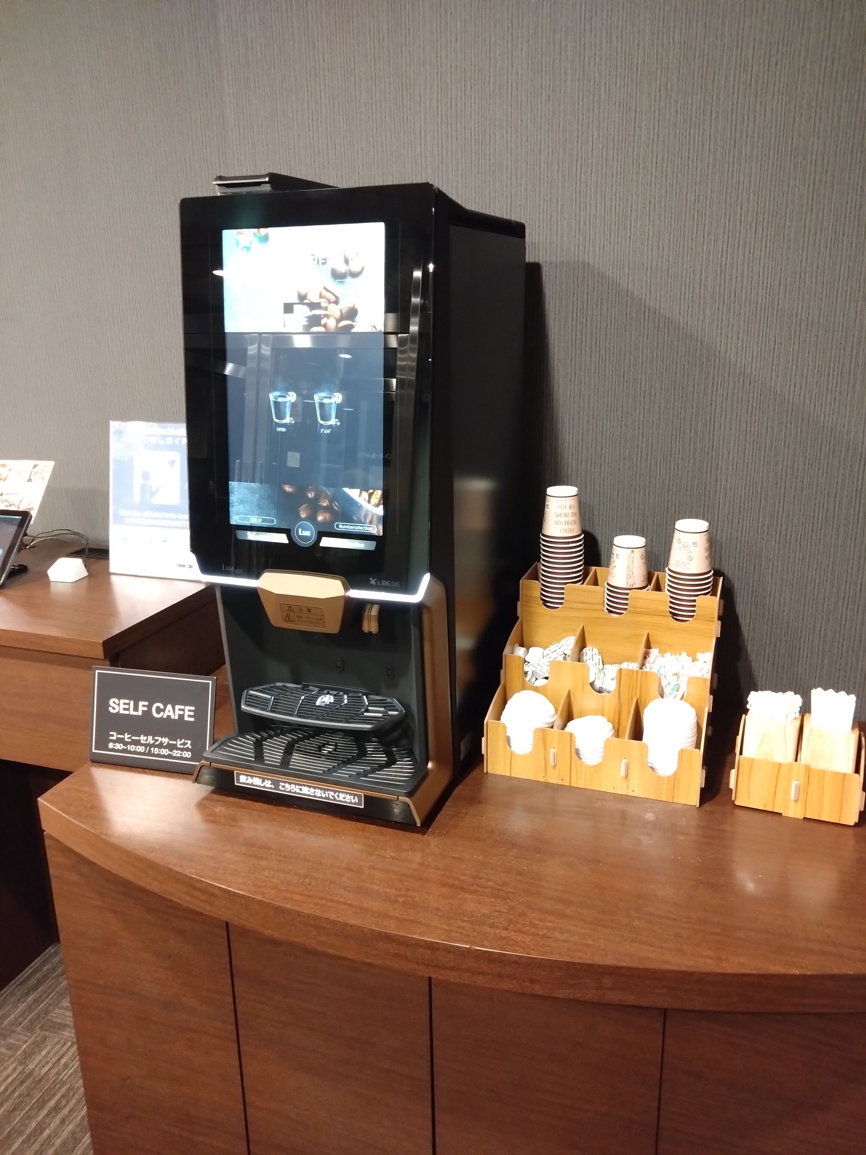 A self-service coffee station with a digital display, coffee pods, and a sign indicating operating hours.