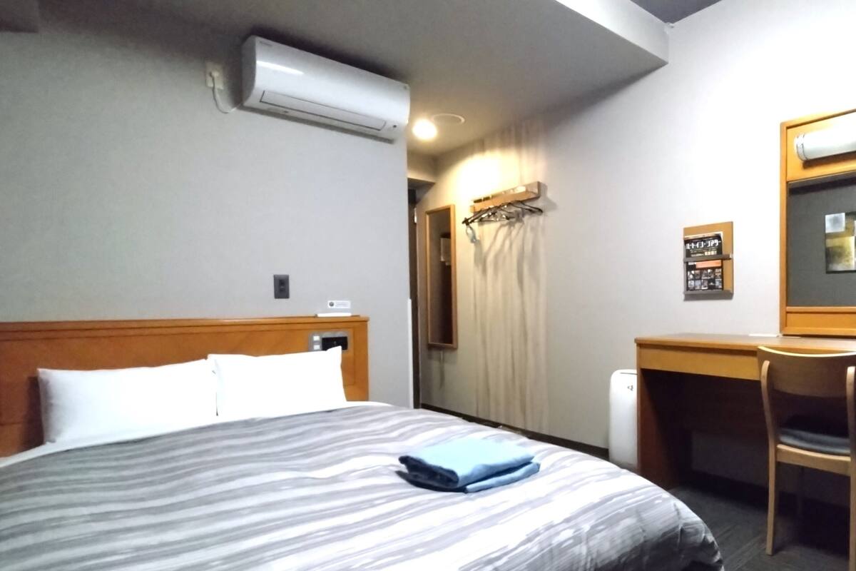A hotel room with a bed, a desk, a chair, an air conditioning unit, and a mirror.