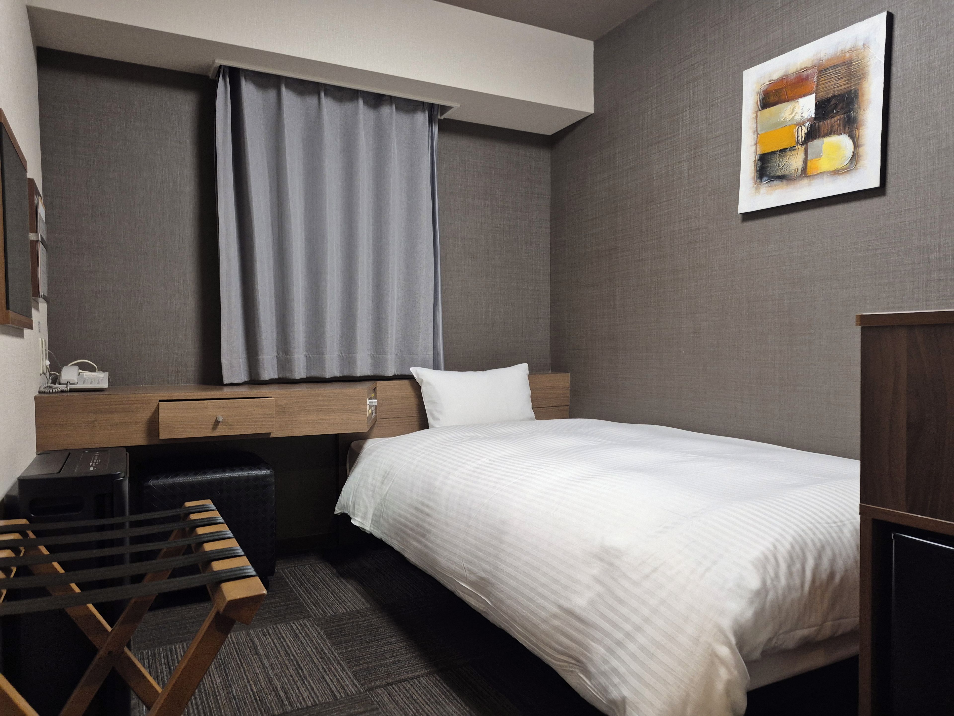A hotel room with a bed, a desk, a chair, a small table, a nightstand, a lamp, a painting, and a window with curtains.