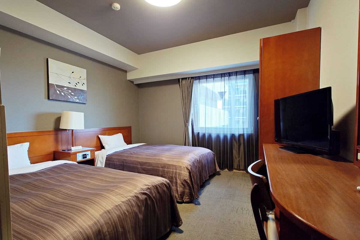 A hotel room with two beds, a desk, a television, and a window with curtains.