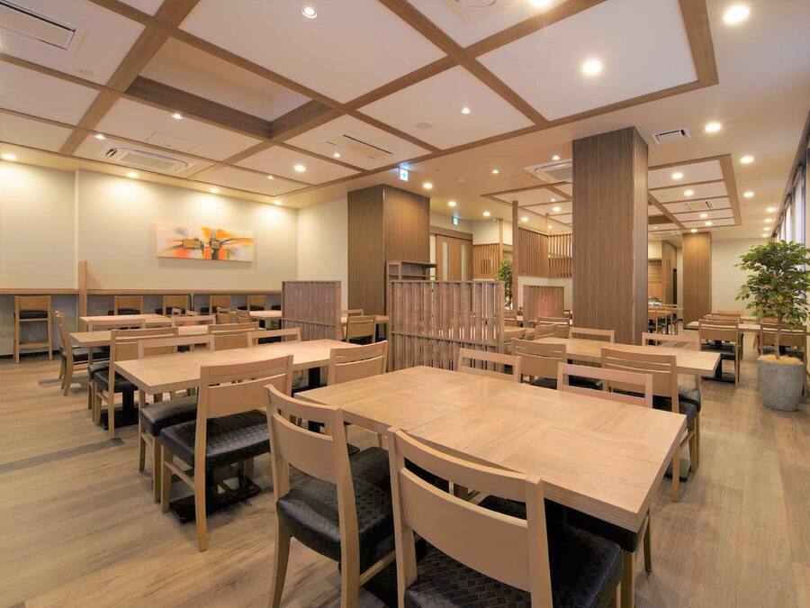 A spacious dining area with wooden tables and chairs, recessed ceiling lighting, and a large potted plant.