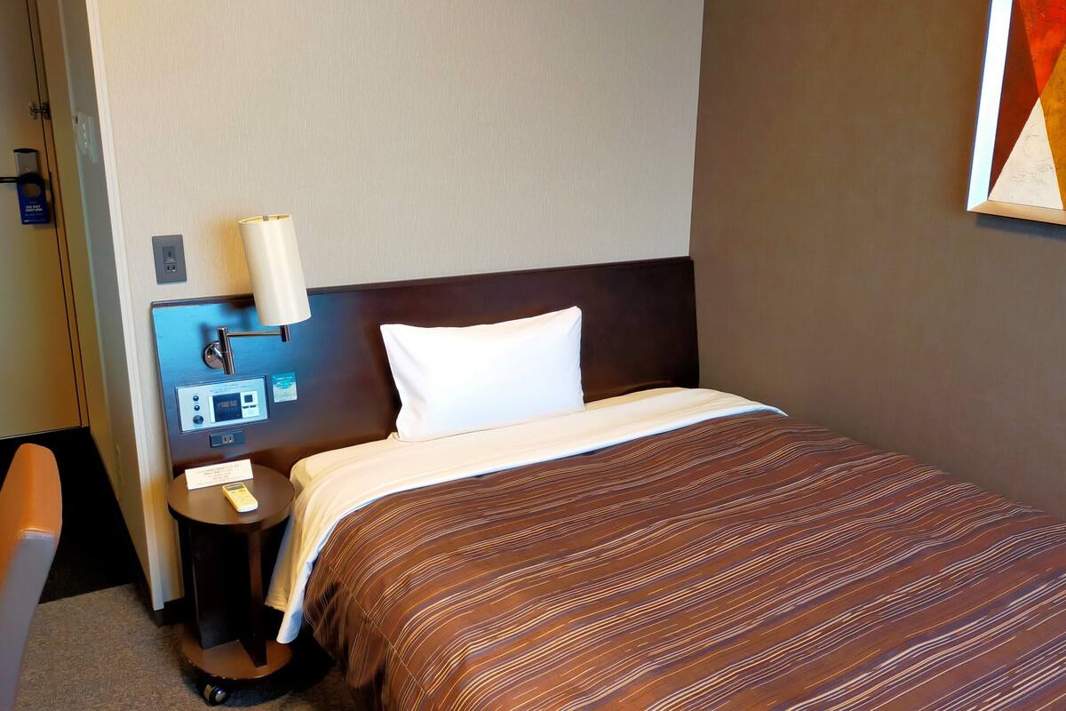 Habitación de hotel con cama, mesita de noche con lámpara, panel de control y un cuadro en la pared.