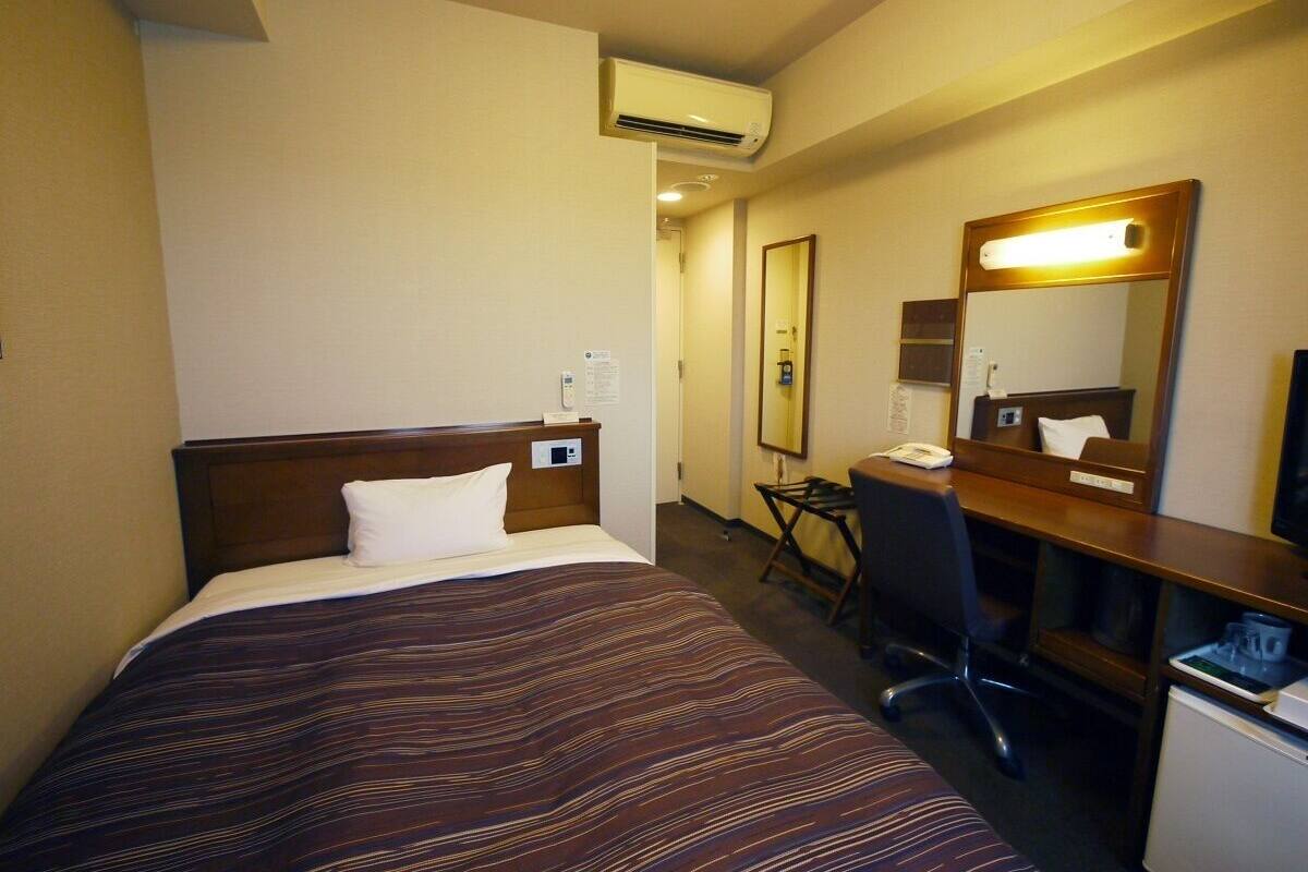 Habitación de hotel con cama, escritorio, silla, espejo y aire acondicionado.
