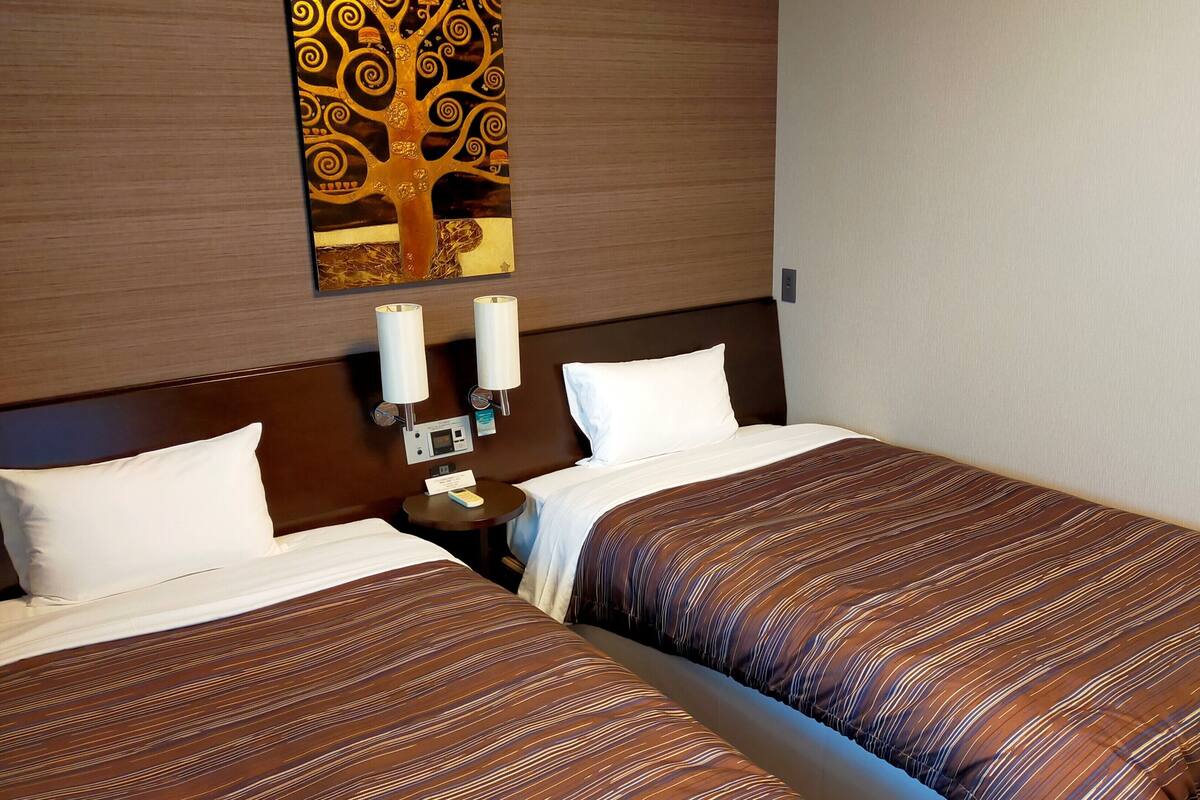 Habitación de hotel con dos camas, una mesita de noche, una lámpara de pared y una obra de arte con un árbol decorativo.
