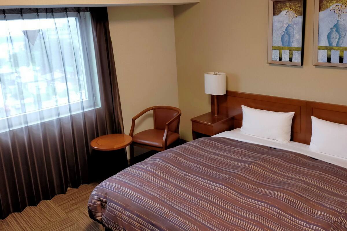 Habitación de hotel con una cama grande, una silla, una mesita de noche con lámpara y una ventana con cortinas.