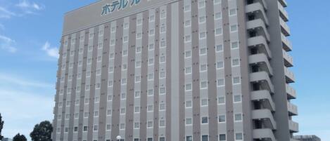 Un hotel de varios pisos con fachada uniforme, un letrero llamativo en la parte superior y un estacionamiento con varios coches.