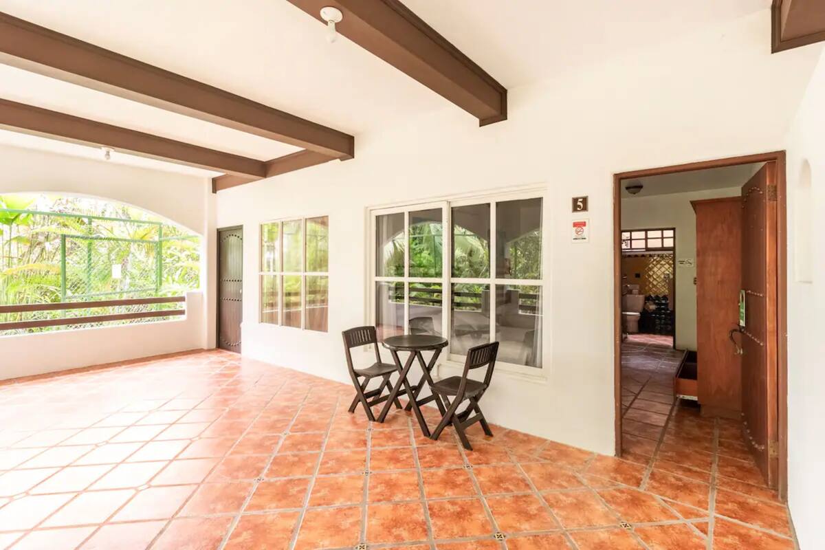 A spacious balcony with a tiled floor, a table with chairs, and large windows offering a view of greenery.