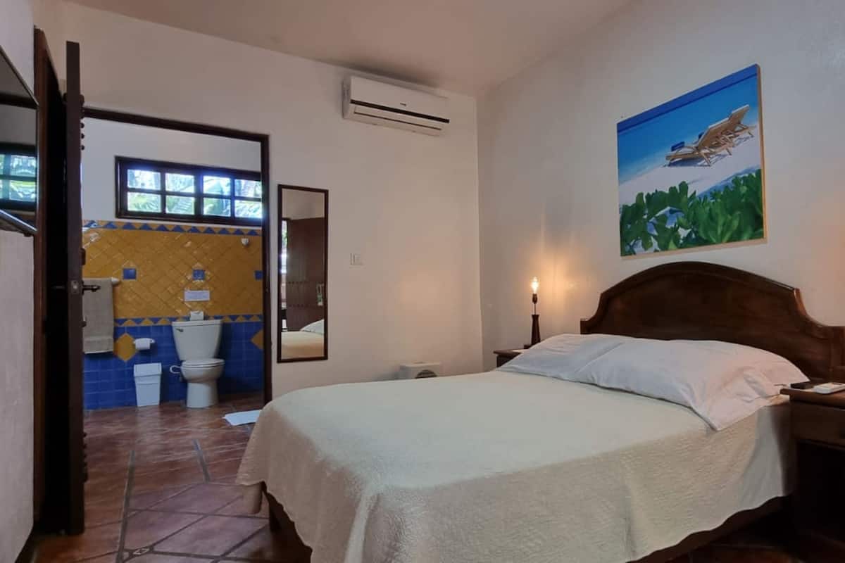A bedroom with a bed, bedside tables, a nightstand, a wall-mounted TV, a painting, and an air conditioning unit.