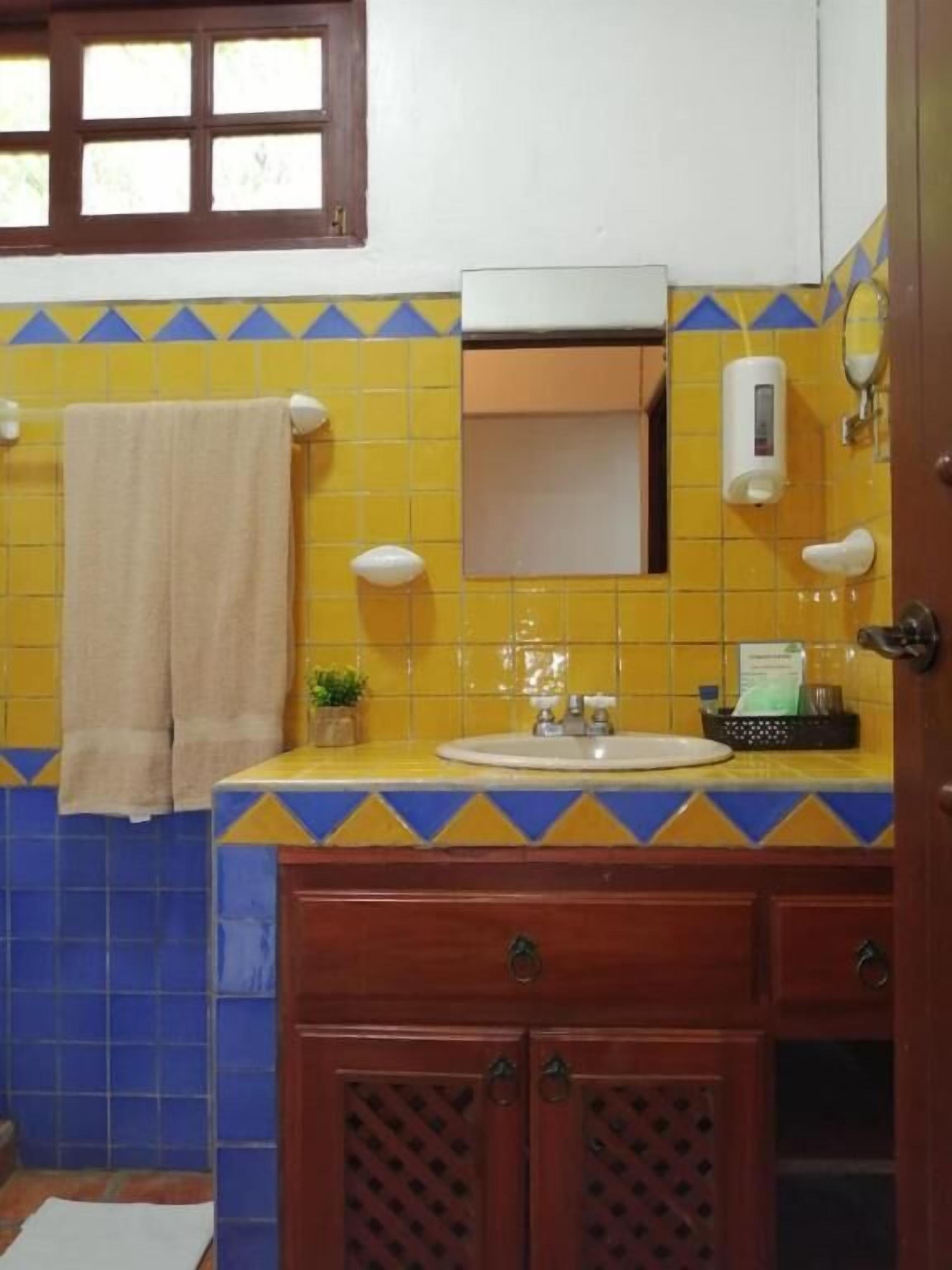 Un bagno con piastrelle gialle e blu, un lavandino con uno specchio sopra, un asciugamano appeso al muro e un dispenser di sapone.