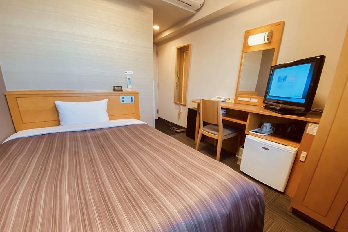 A hotel room with a bed, a desk with a computer, a small refrigerator, and a mirror.