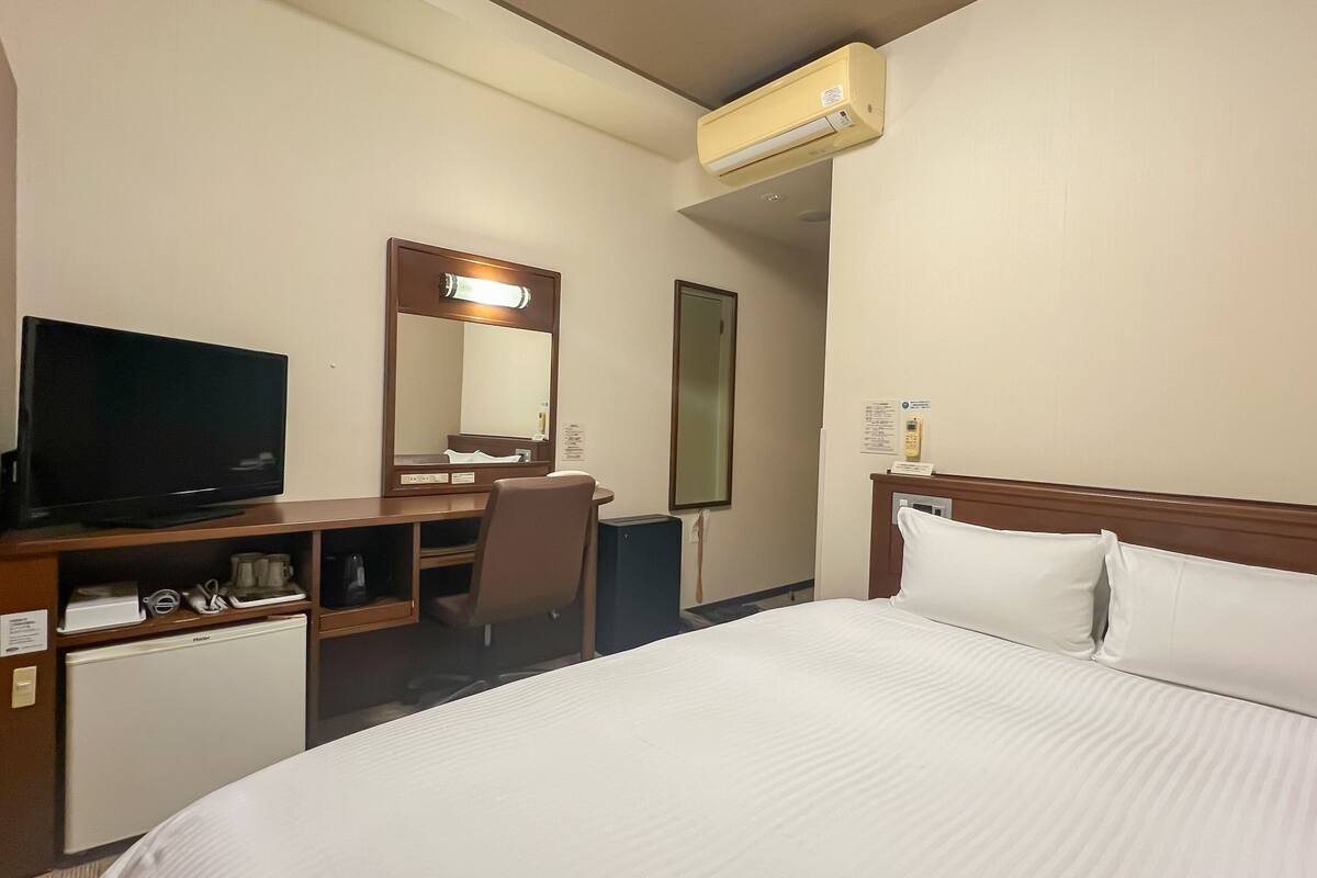 A hotel room with a bed, a television, a desk with a chair, a mirror, and an air conditioning unit.