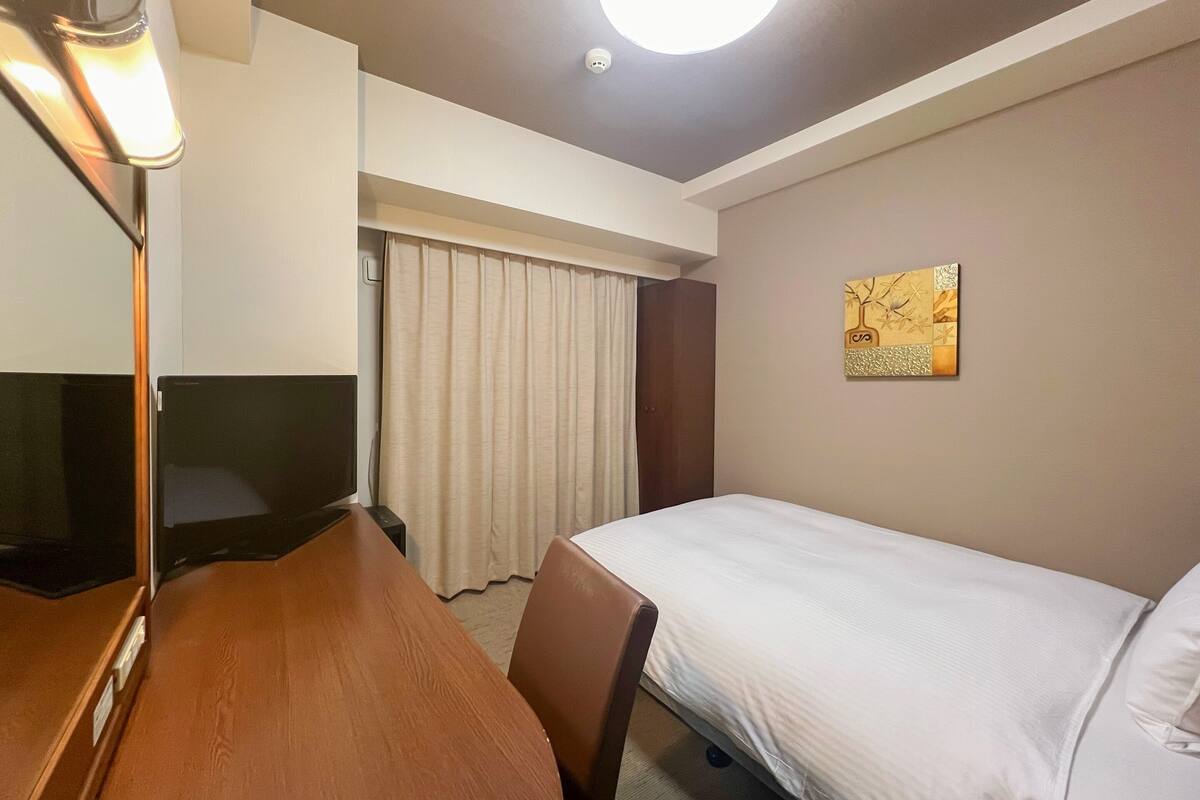 A hotel room with a bed, a desk with a chair, a television, a curtain, and a ceiling light.