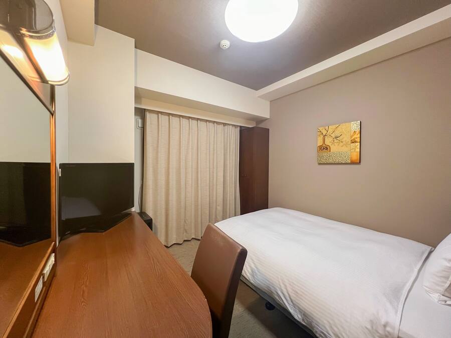 Une chambre d’hôtel comprenant un lit, un bureau avec une chaise, une télévision, un rideau et un luminaire au plafond.