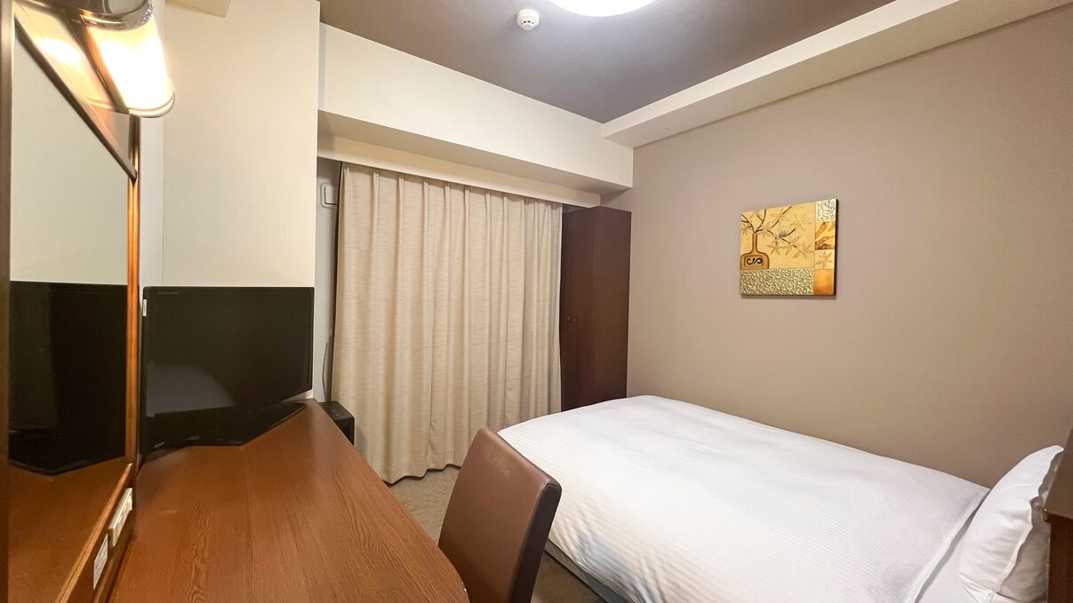 A hotel room with a bed, a desk with a television, a chair, a window with curtains, and a wall-mounted artwork.