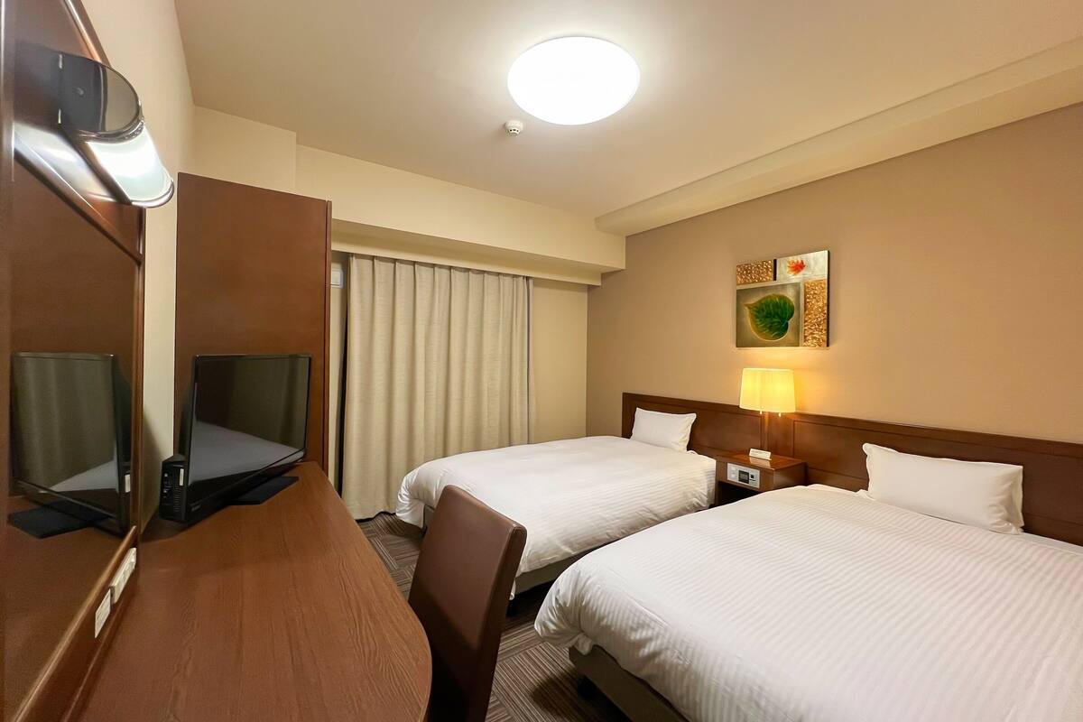 A hotel room with two beds, a desk, a chair, a desk lamp, and a wall-mounted artwork.