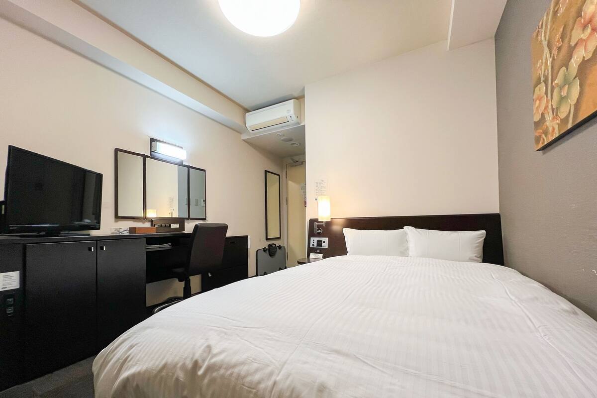 A hotel room with a large bed, a desk with a computer, a television, and an air conditioning unit.