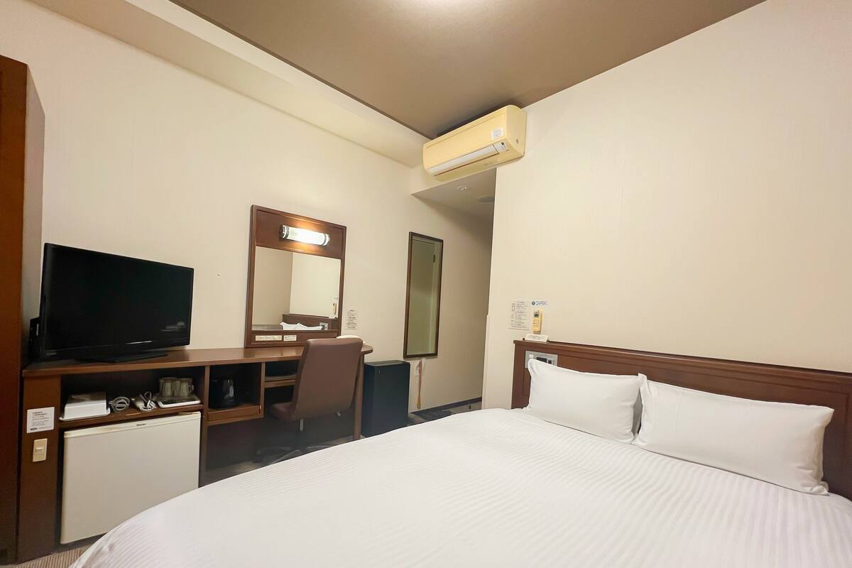 A hotel room with a bed, a desk with a television, a chair, a mirror, and an air conditioning unit.
