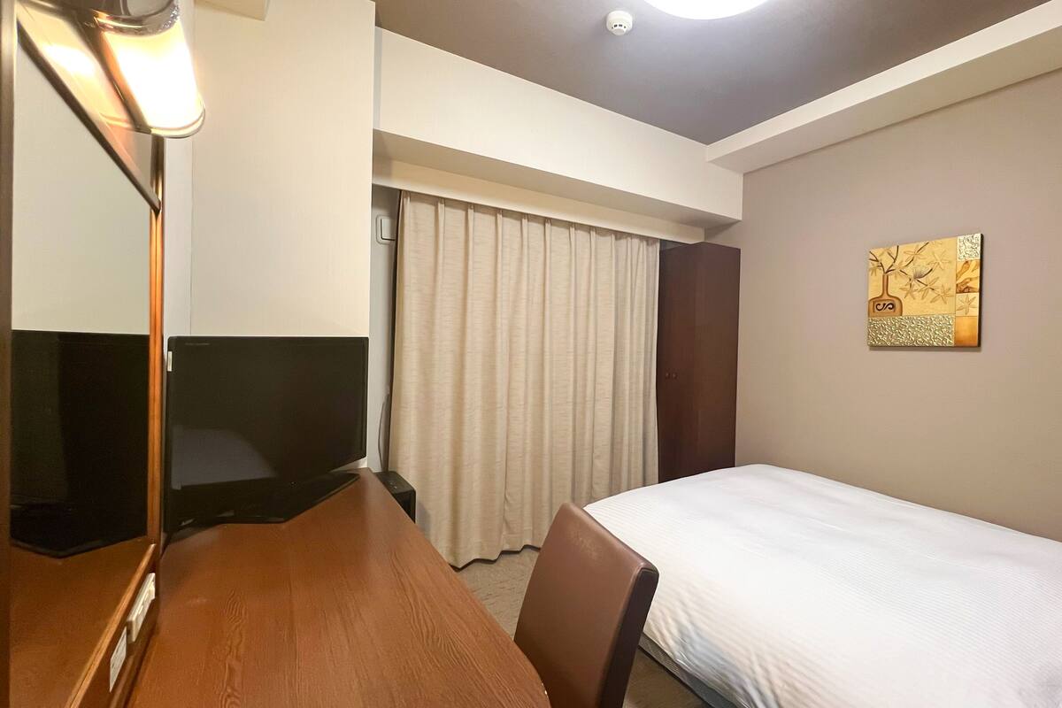 A hotel room with a bed, a desk, a television, a window with curtains, and a wall-mounted artwork.