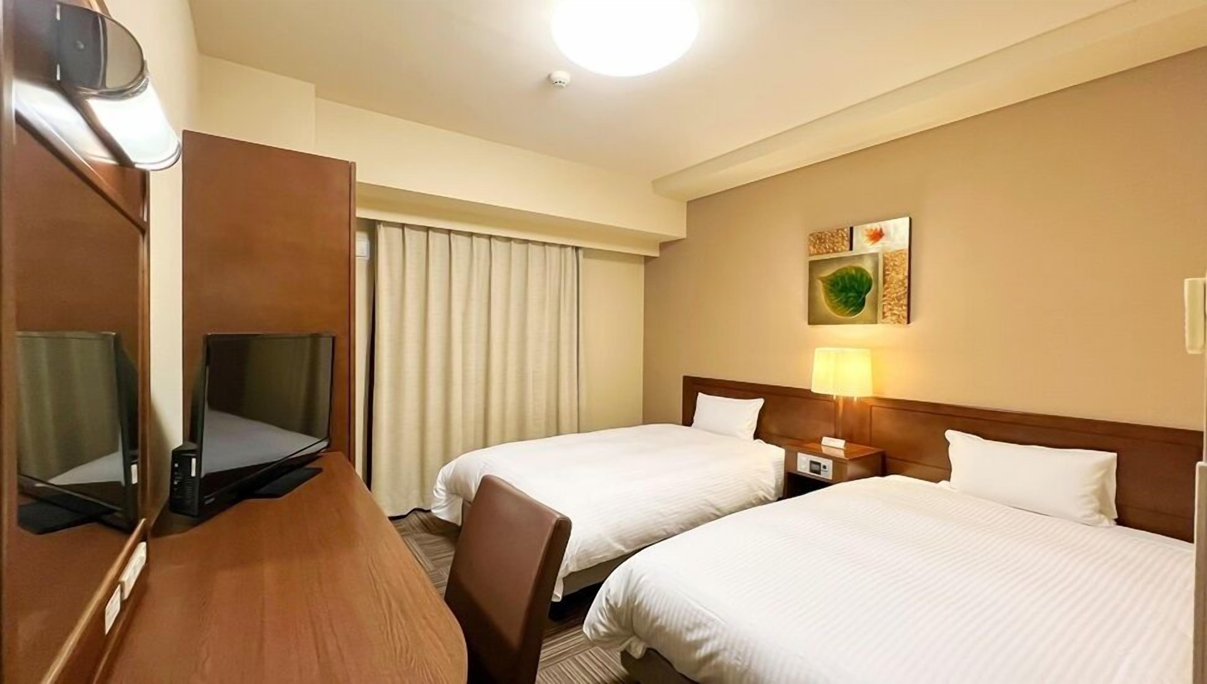 A hotel room with two beds, a desk with a television, a lamp, and a window with curtains.