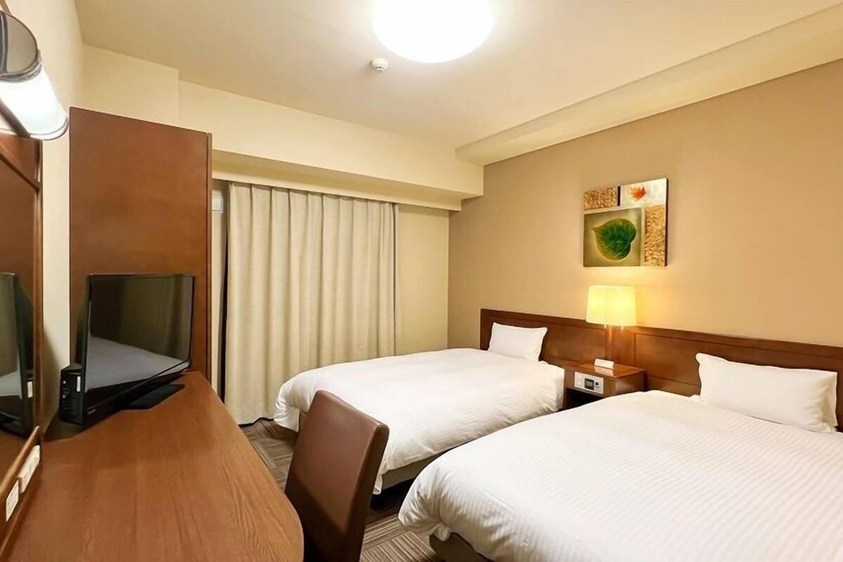 A hotel room with two beds, a desk with a television, a lamp, and a window with curtains.