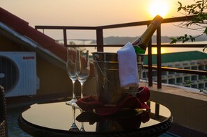 Une table avec une bouteille de champagne, deux verres et un seau recouvert d’un linge, sur un balcon donnant sur un coucher de soleil et un pont.