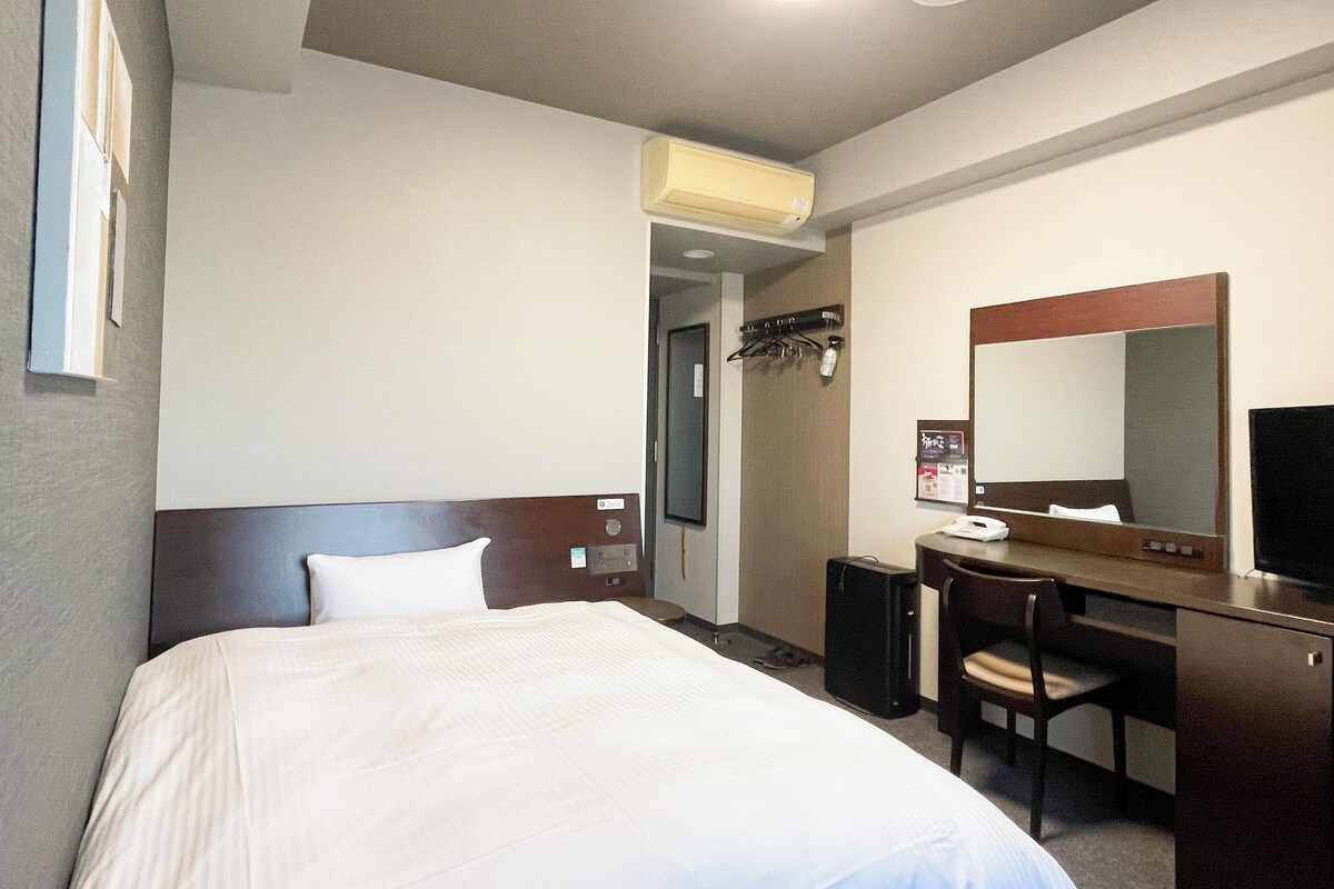 A hotel room with a bed, a desk, a chair, a mirror, and a wall-mounted air conditioner.