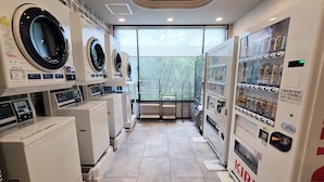 A hotel laundry room with multiple washing machines and dryers, a vending machine, and a window with a view of bamboo outside. - Hotel Route-Inn Odate Eki Minami (Odate)