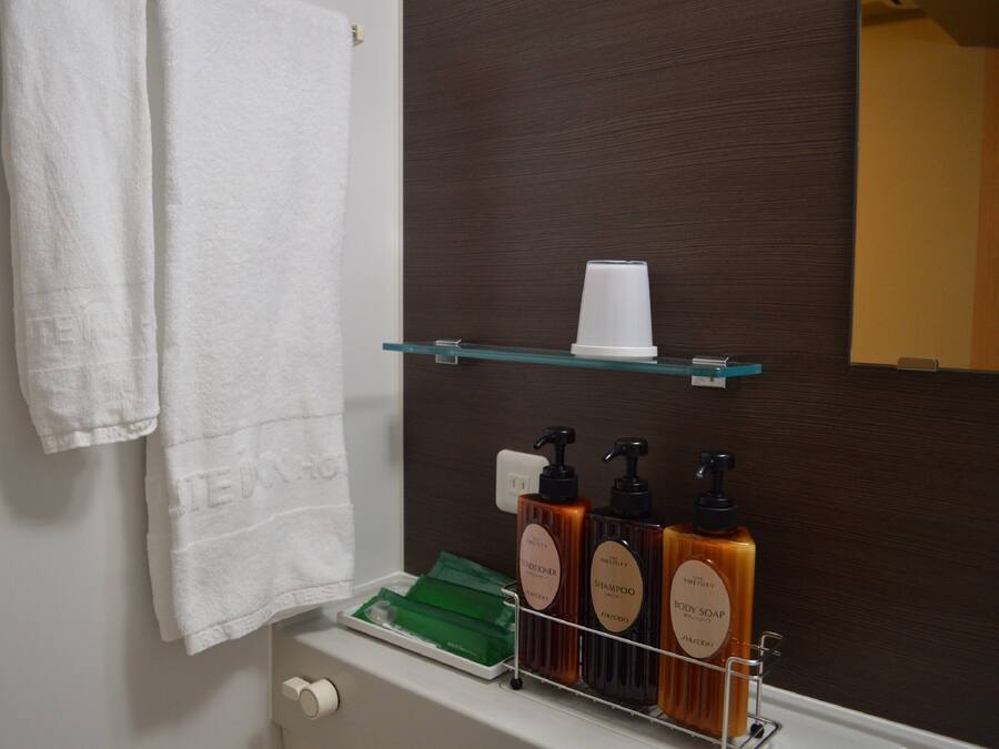Um banheiro com uma toalha branca pendurada na parede, uma prateleira de vidro com uma xícara e três frascos de sabonete líquido.