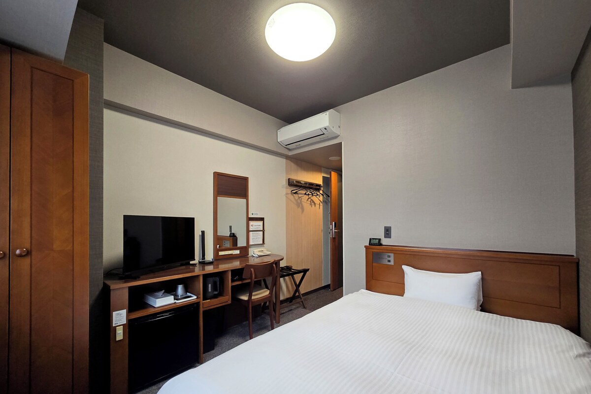 A hotel room with a bed, a desk with a TV, a mirror, a coat rack, and an air conditioning unit.