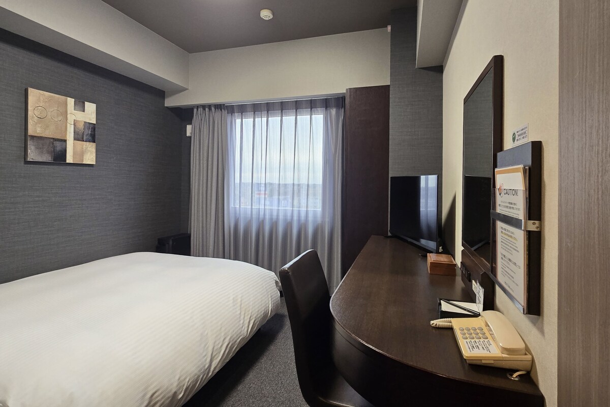 A hotel room with a bed, a desk with a telephone, a television, and a window with curtains.
