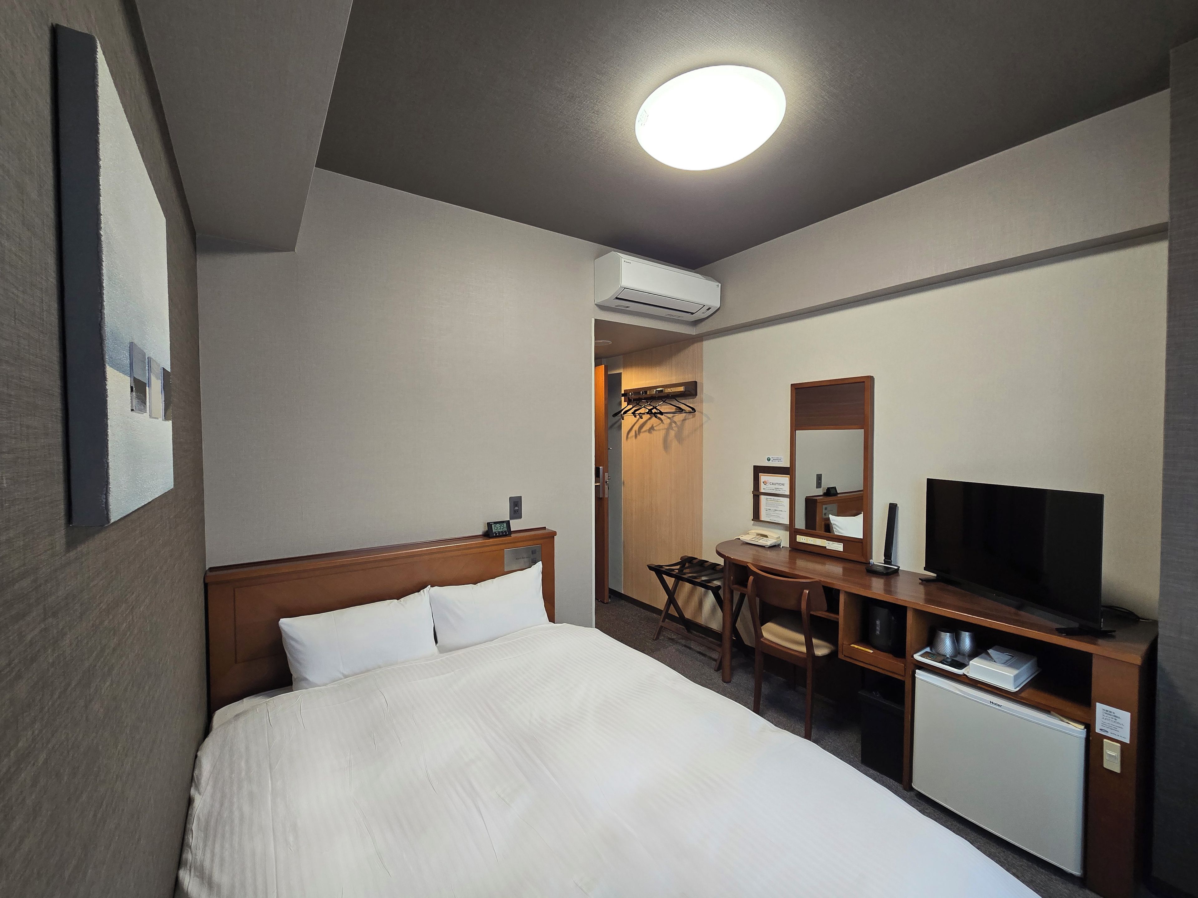 A hotel room with a bed, a desk with a television, a mini-fridge, and a mirror.