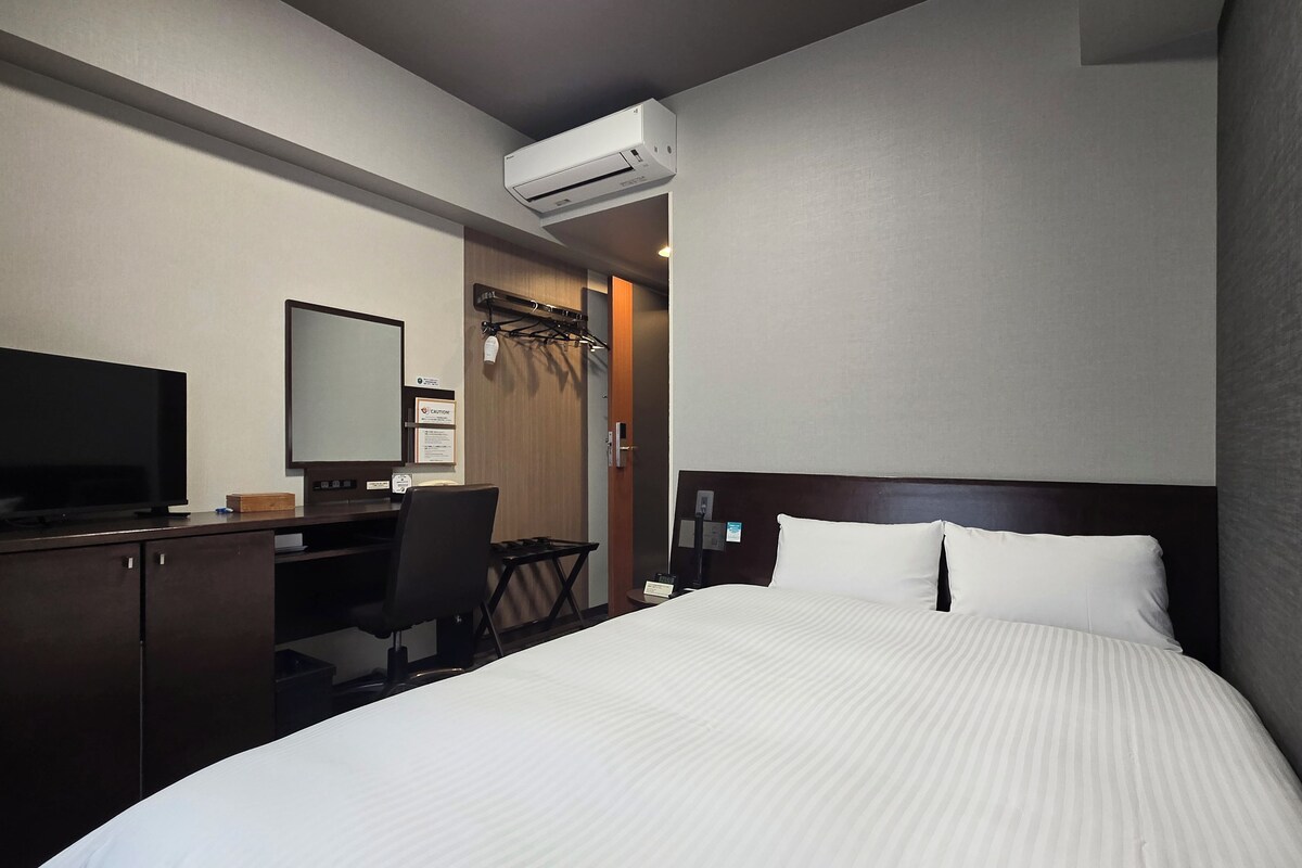 A hotel room with a bed, a desk with a computer, a television, and an air conditioning unit.