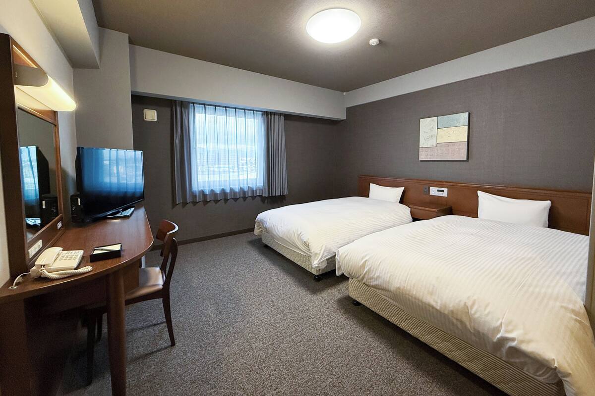A hotel room with two beds, a desk with a telephone, a television, and a window with curtains.