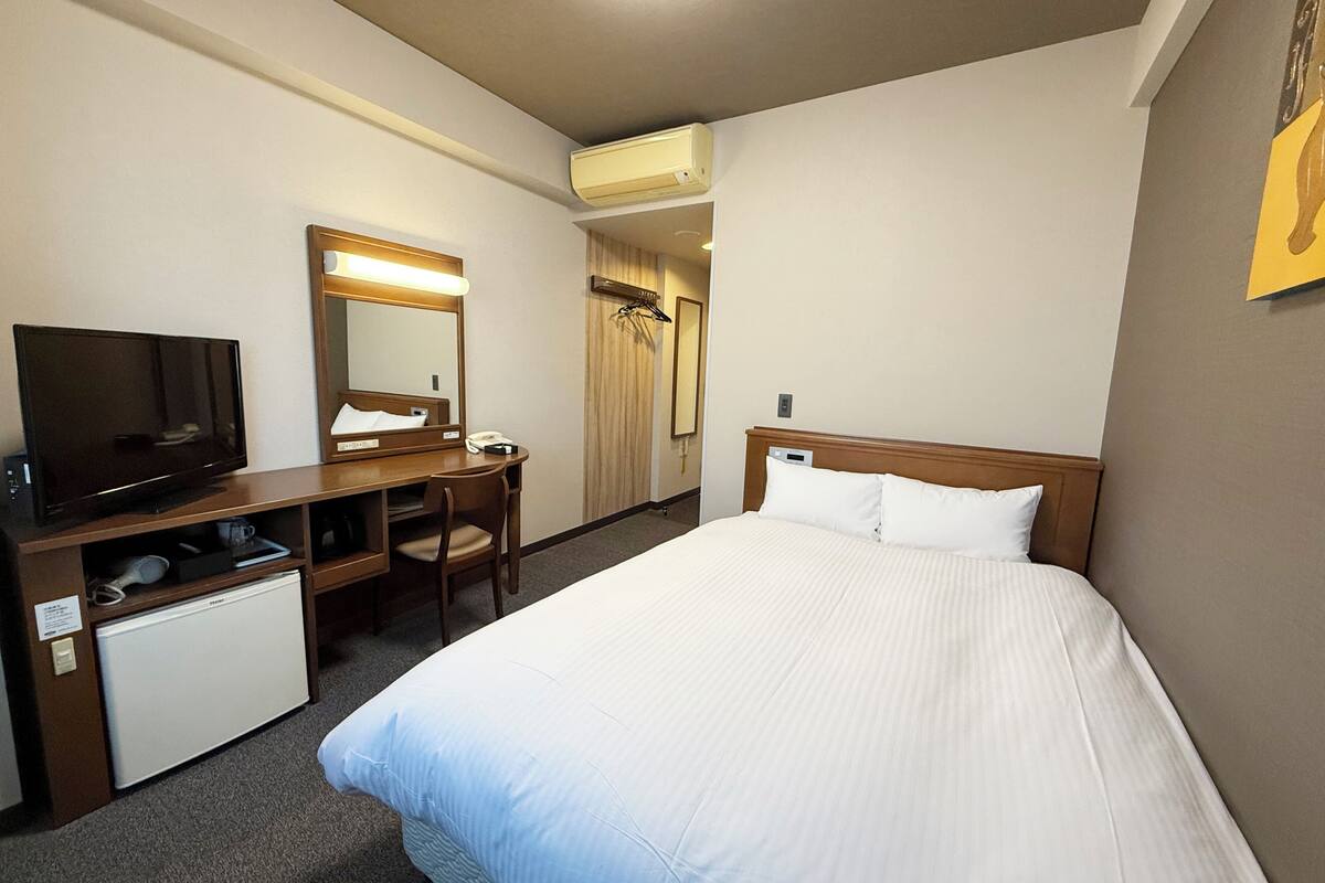 A hotel room with a bed, a desk with a television, a mirror, and a small refrigerator.