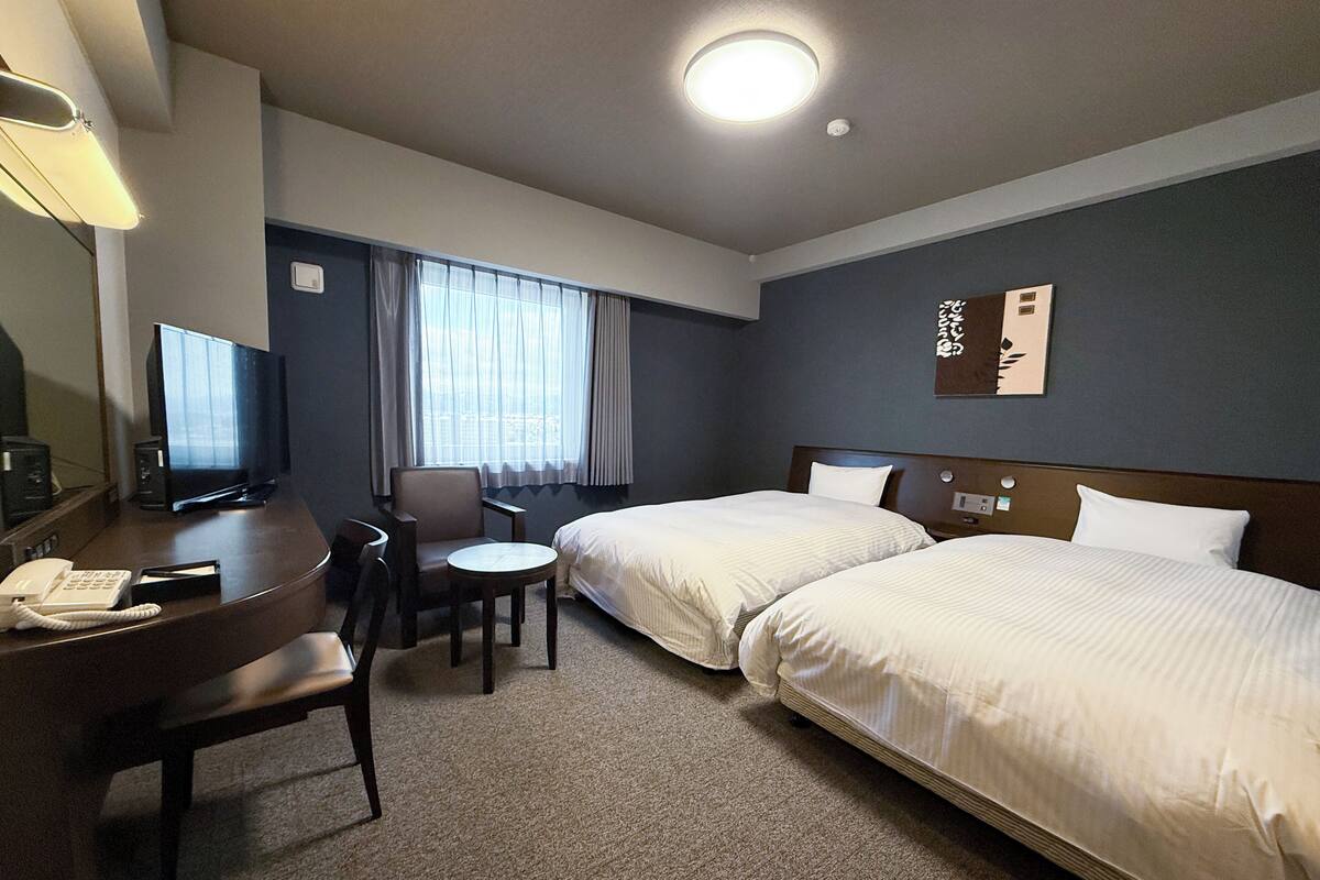 A hotel room with two beds, a desk with a telephone, a chair, a small table, and a television.